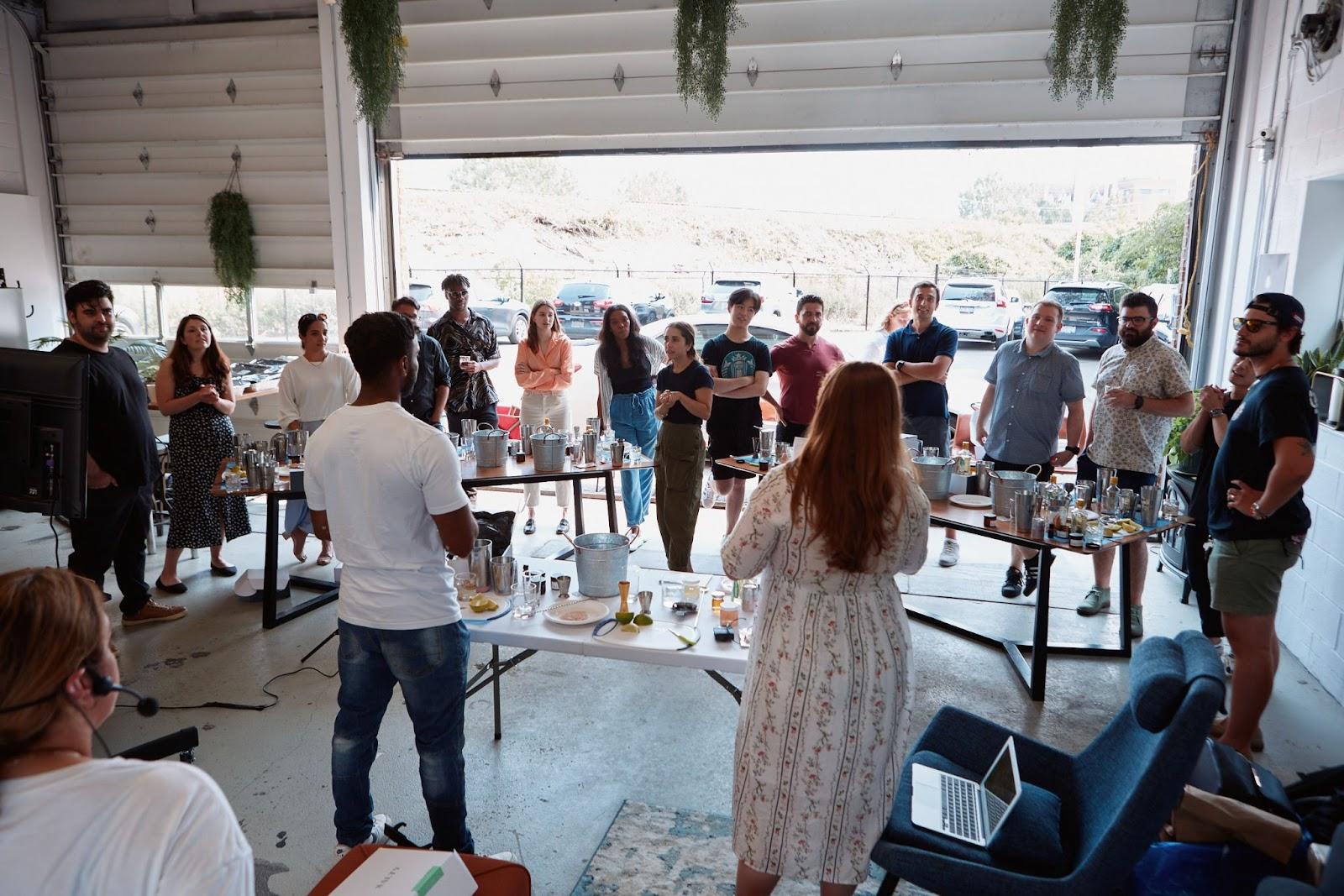 Group of people gathered indoors around tables with cocktail-making supplies, listening to a presentation from two instructors.