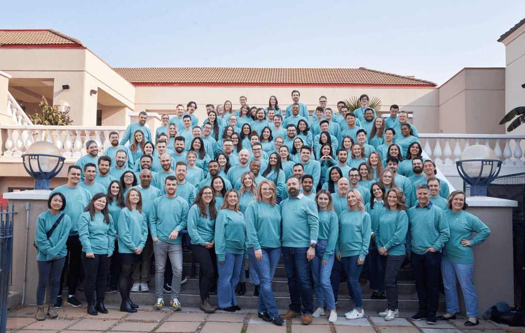 A large group of Coconut Software temmembers, all wearing matching teal sweatshirts, standing on outdoor steps in front of a building with red-tiled roofs, posing for a team photo.