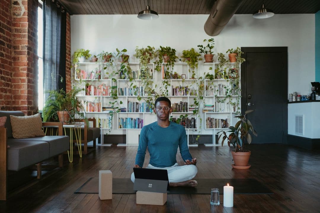 A man sits cross-legged in a well-lit room with plants and bookshelves, meditating on a yoga mat, with a laptop, candle, and glass of water nearby.