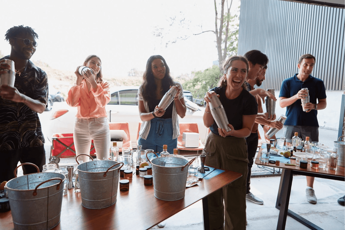 Group of people at an outdoor workshop smiling and shaking cocktail shakers, with tables set up with ingredients and tools.