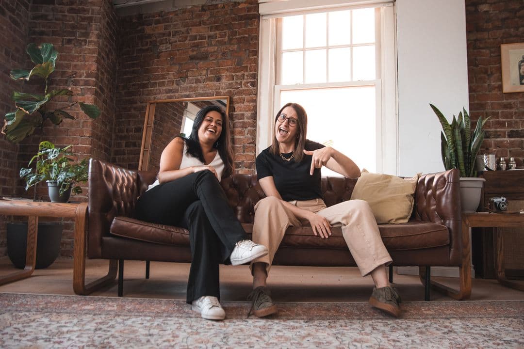 Wavy founders sitting on a leather couch in a brick-walled room, laughing and talking.